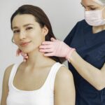 A medical practitioner in scrub suit performs a neck examination on a smiling woman in a clinic.